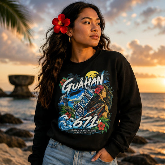 A woman stands on a beach at sunset wearing a black crewneck sweatshirt with a colorful graphic print featuring the text 'GUAMAN 672' and tropical artwork. She has a red flower in her hair and is wearing light blue jeans.